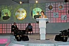 First Lady Laura Bush speaking at opening ceremony, with Attorney General Gonzales and Assistant Attorney General Schofield seated in background.