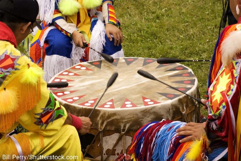 Stock photo of a Native American celebration, with tribal members in traditional dress seated around a drum.